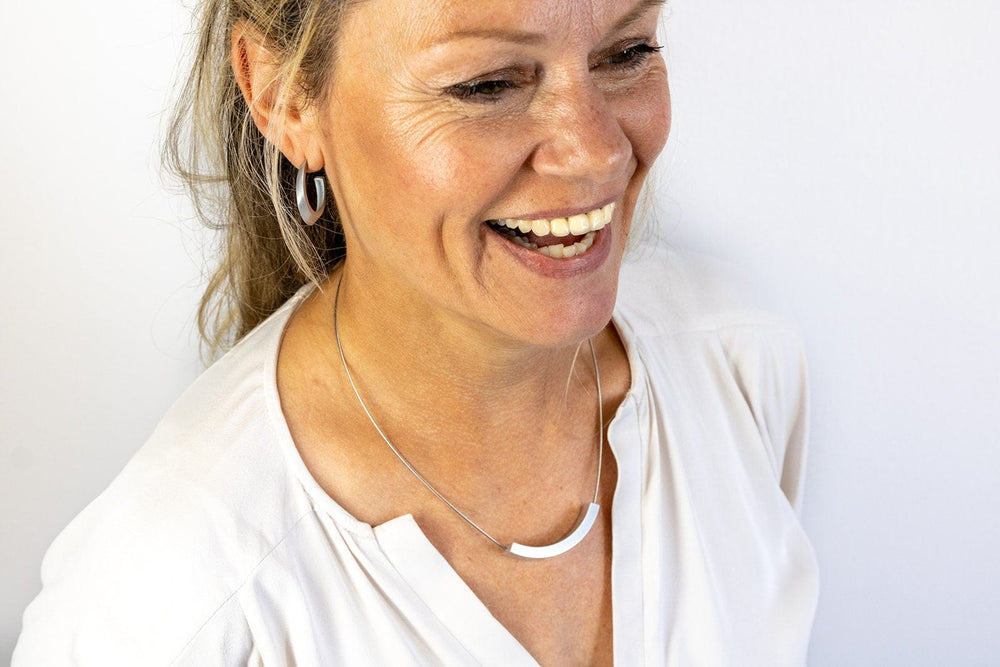 Een lachende vrouw in een lichte blouse draagt bijpassende geometrische oorbellen en een CLIC Creations Collier Bogen Goud|Geel C74G. Haar lichtbruine haar is naar achteren gebonden en ze staat tegen een effen witte achtergrond.