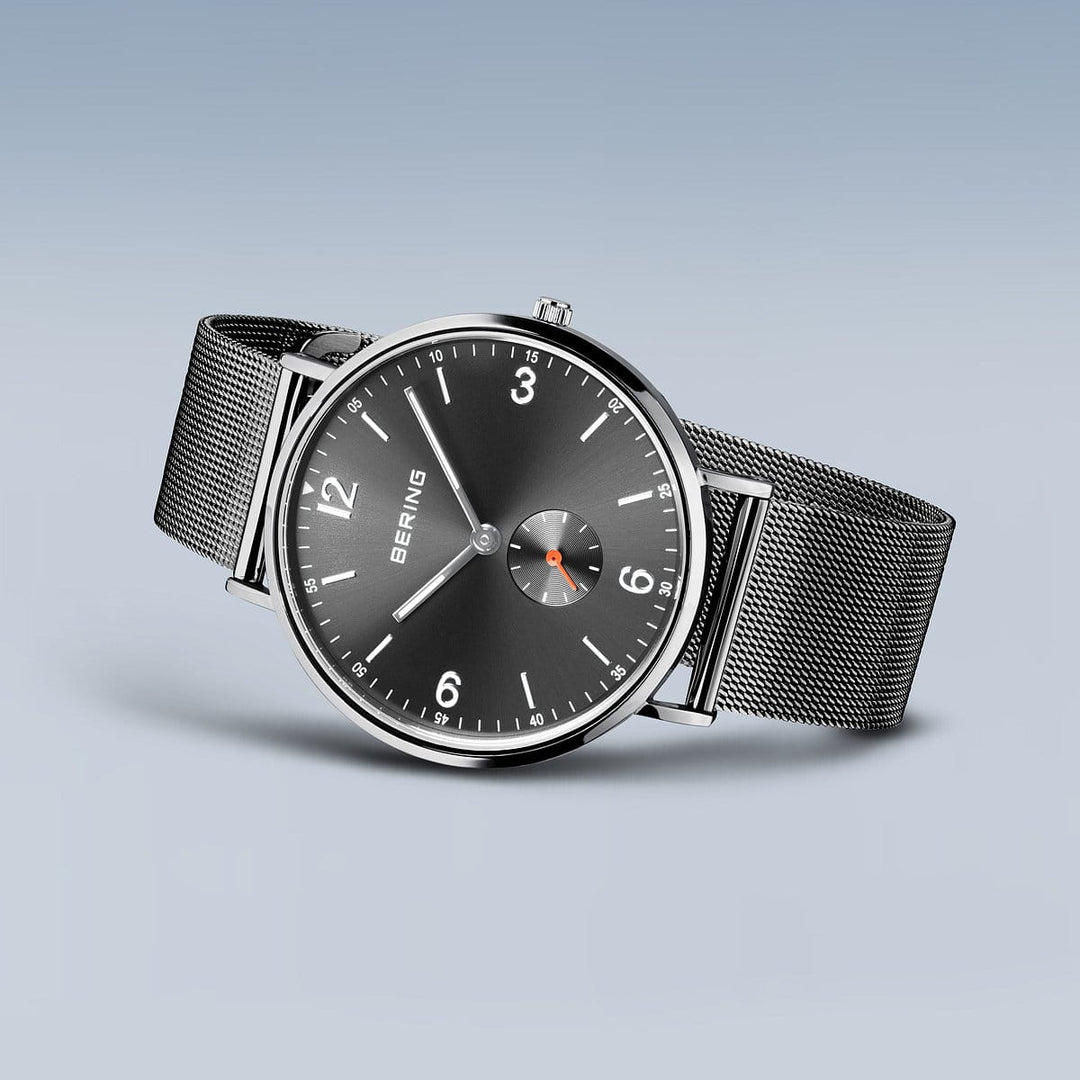 De Bering heren horloge grijs 14040-377 heeft een gestroomlijnd zilverkleurig ontwerp, een zwarte wijzerplaat met minimalistische witte markeringen, een fijnmazige band en een elegant kwartsuurwerk - afgebeeld tegen een blauwgrijze achtergrond met kleurverloop.