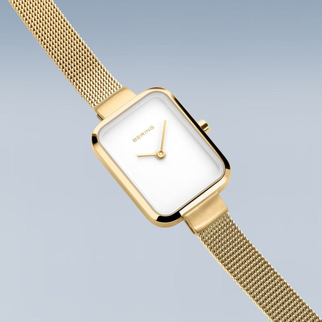 Het Bering dameshorloge 14520-334 met goudkleurige mesh band, roestvrijstalen kast en rechthoekige witte wijzerplaat zonder cijfers wordt getoond op een lichtblauwe achtergrond.