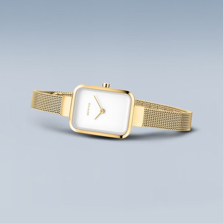 De Bering 14520-334 dameshorloge heeft een minimalistisch goudkleurig ontwerp met een vierkante witte wijzerplaat, roestvrijstalen kast, goudkleurige mesh band en eenvoudige wijzers op een lichtgrijsblauwe achtergrond.