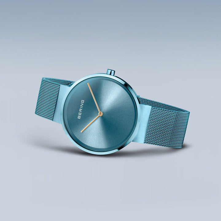 De Bering 14539-388 is een strak minimalistisch horloge met een blauwe mesh band, bijpassende blauwe wijzerplaat, zilveren uur- en minutenwijzers en saffierglas. Het gestroomlijnde ontwerp toont de merknaam BERING op de wijzerplaat tegen een verloop van grijze en blauwe tinten.