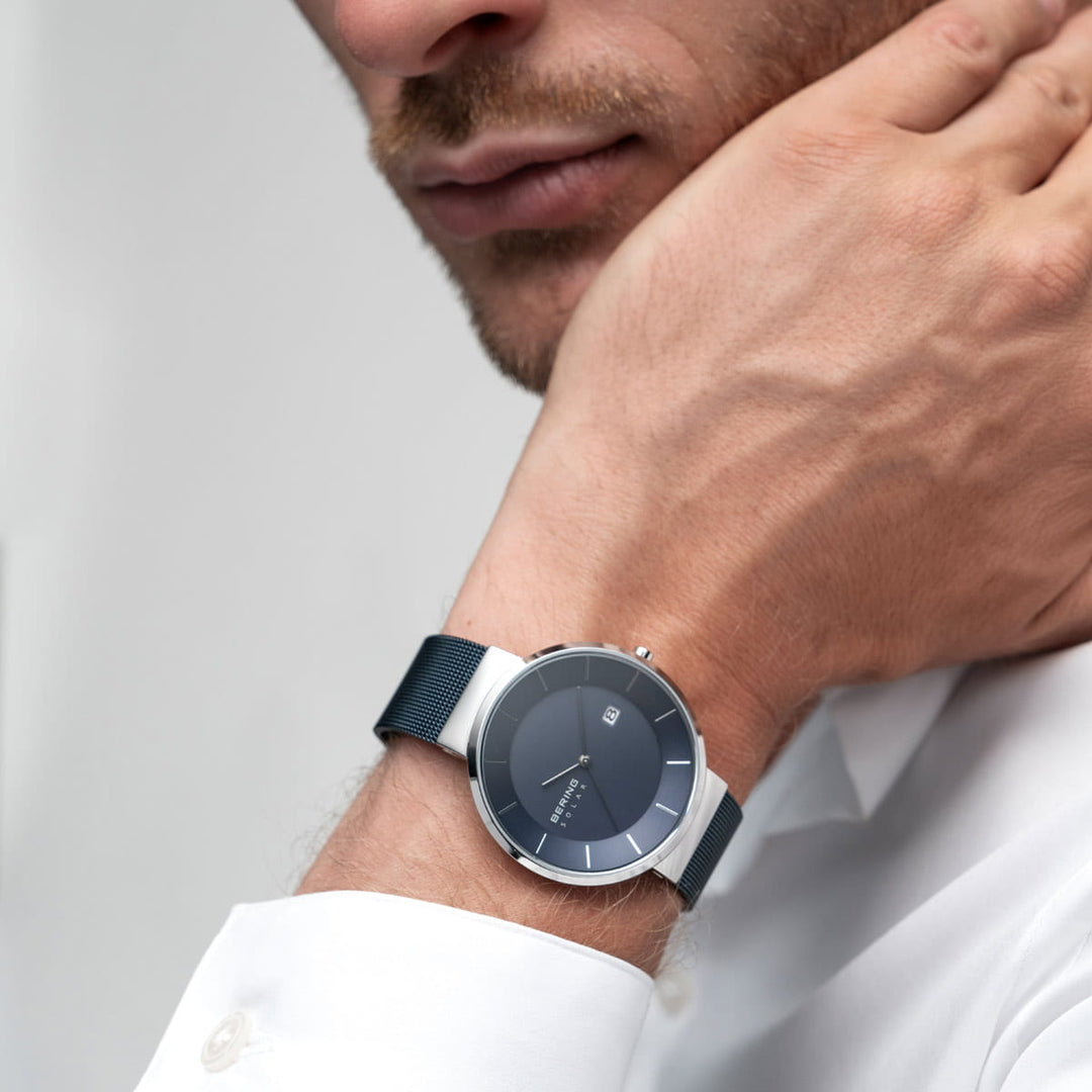 Een man in een wit overhemd, in een minimalistische setting, rust zijn kin op zijn hand, en toont het Bering horloge 14639-307 met een donkere wijzerplaat en mesh band. Het horloge is elegant voorzien van Saffierglas en solar-uurwerk technologie.