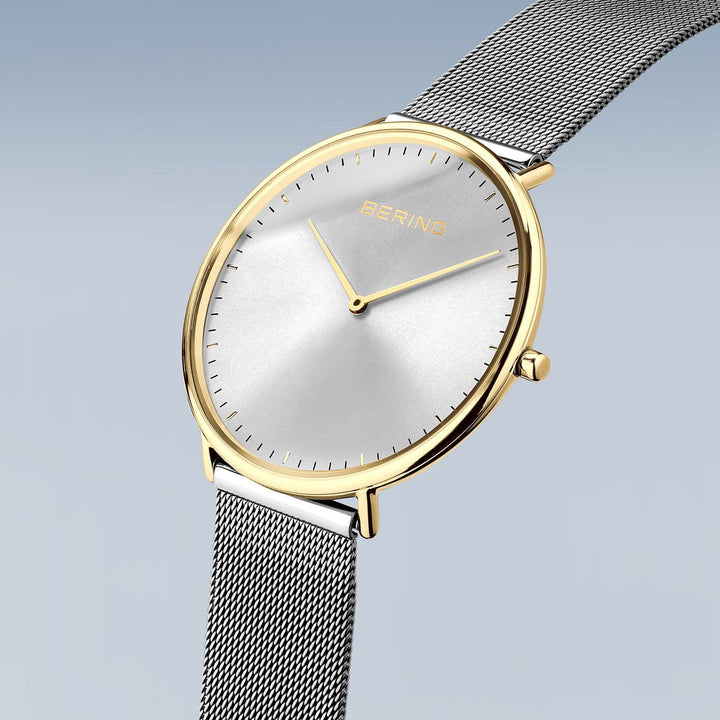 Het Bering 15739-010 Dameshorloge toont tijdloze elegantie met zijn minimalistische design, met een ronde, zilveren kast, zilveren wijzerplaat, dunne zwarte uurmarkeringen, twee gouden wijzers en een mesh-band op een lichtblauwe achtergrond.