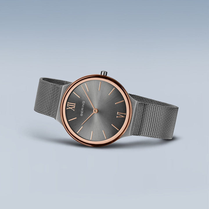 Het Bering dameshorloge 18434-369 heeft een roségouden kast, grijze wijzerplaat met Romeinse cijfers en grijze mesh band. Het is voorzien van duurzaam saffierglas en wordt elegant weergegeven op een lichtblauwe achtergrond.