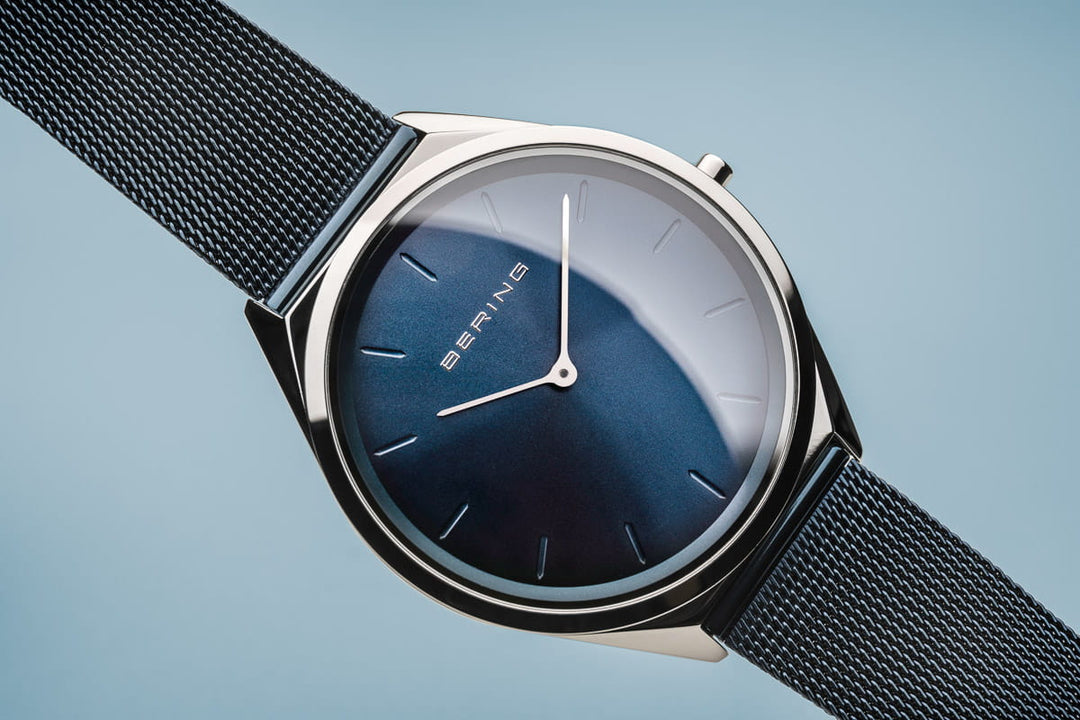 Het Bering unisex horloge (17039-307) heeft een strak minimalistisch design, met een donkerblauwe wijzerplaat met dunne zilveren uurmarkeringen onder saffierglas. Het heeft een quartz uurwerk in een slanke zilveren kast en een fijne zwarte mesh band tegen een lichtblauwe achtergrond.