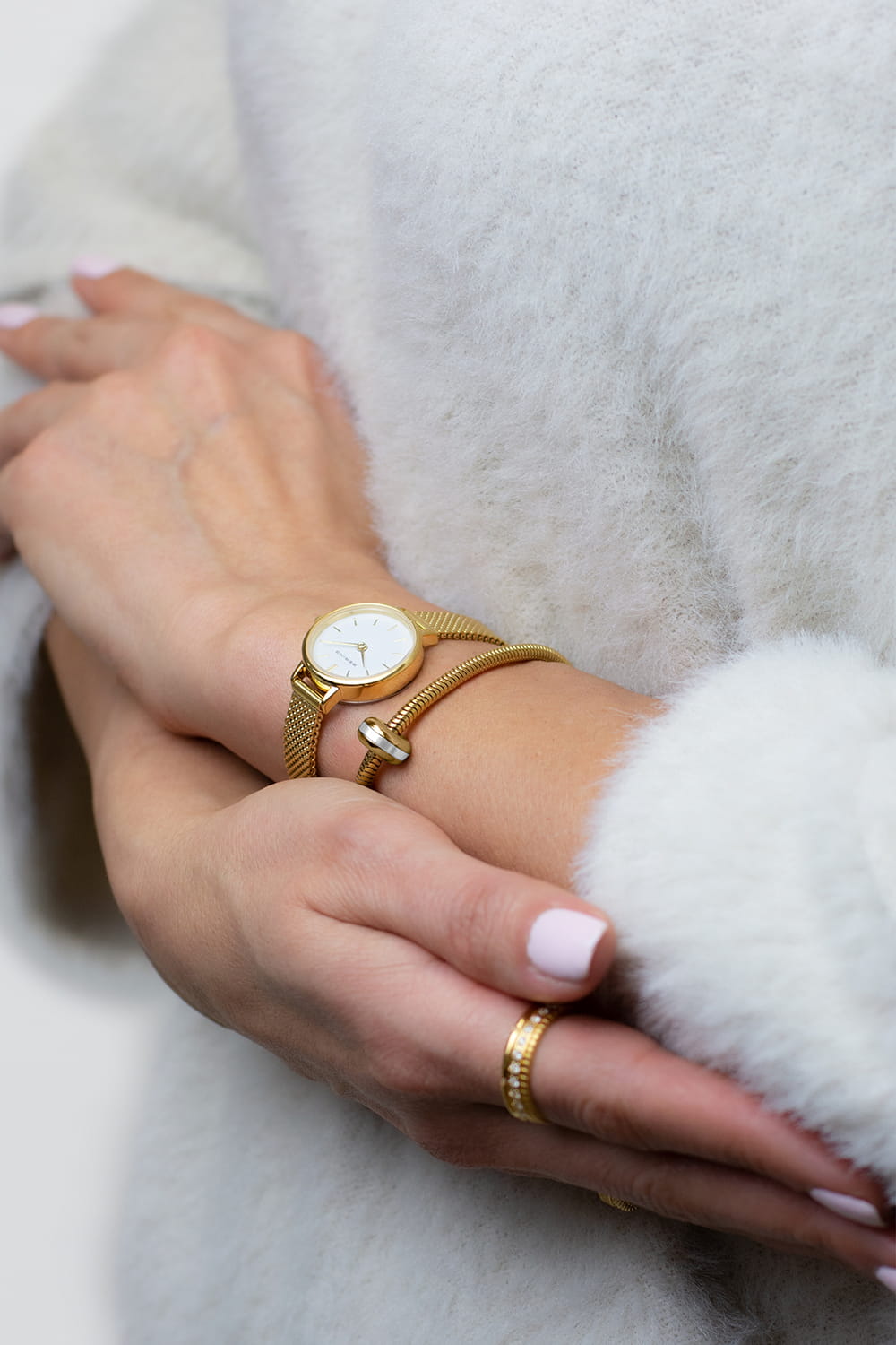 Close-up van een persoon die een Bering 11022-334-Lovely-1-GWP190 draagt met een wit gezicht en gouden armband, met Quartz uurwerk. Ze hebben lichtroze nagellak, dragen een fuzzy witte trui en hebben een getextureerde gouden ring aan hun wijsvinger.