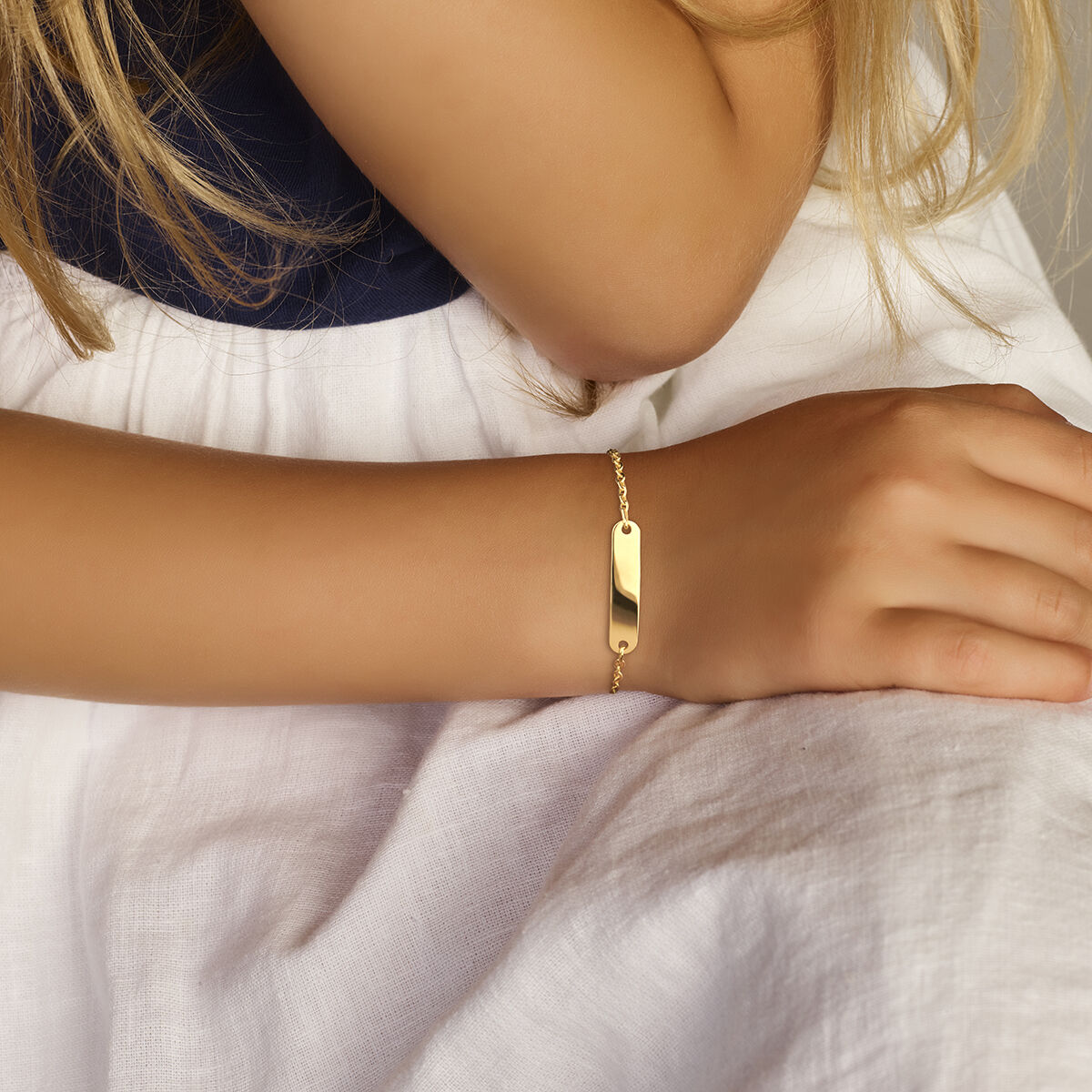 Een close-up van een jonge hand op witte stof, met de Van der Kooij Collectie Baby armband plaat 4,0 mm 9K met een blanco rechthoekige plaat en schakels - een ideaal geboorte geschenk.