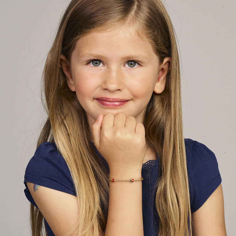 Een jong meisje met lang blond haar en een marineblauw shirt glimlacht naar de camera, haar hand onder haar kin, met om haar pols de Van der Kooij Collectie armband lieveheersbeestje 11-13 cm in 14K geelgoud.