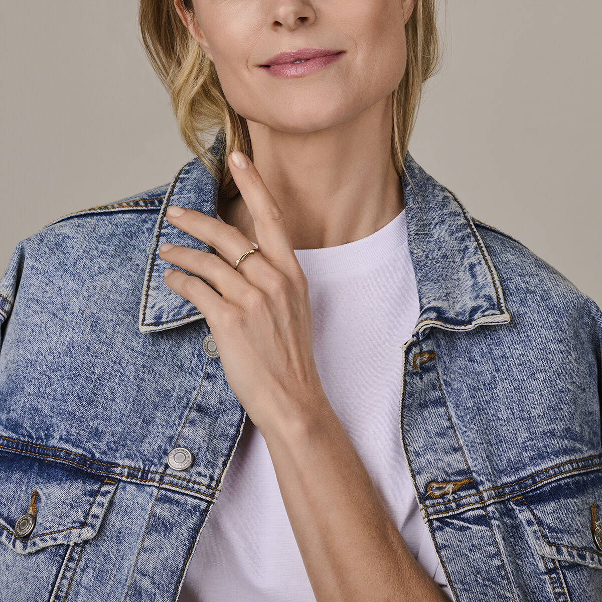 Een vrouw in een blauw spijkerjasje en wit overhemd raakt voorzichtig haar hals aan en onthult een ring 14K geelgoud van Van der Kooij Collectie. Haar glimlach en de zichtbare onderste helft van haar gezicht benadrukken haar tijdloze elegantie.