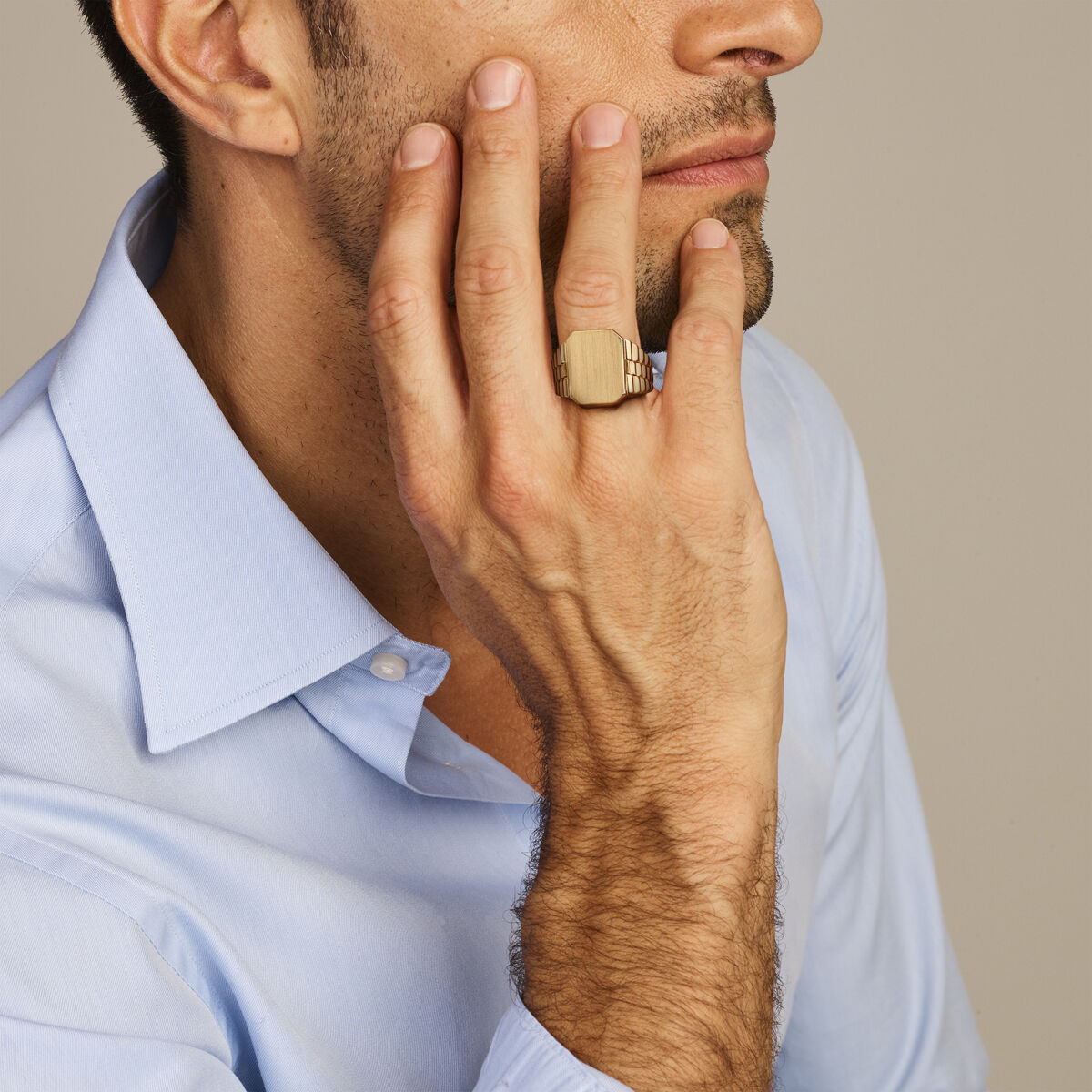 Een man in een lichtblauw overhemd raakt zijn gezicht aan, terwijl hij hetveerring gematteerd 14K geelgoud van Van der Kooij Collectie laat zien. Zijn bedachtzame pose en de neutrale achtergrond benadrukken de elegante eenvoud van de ring.