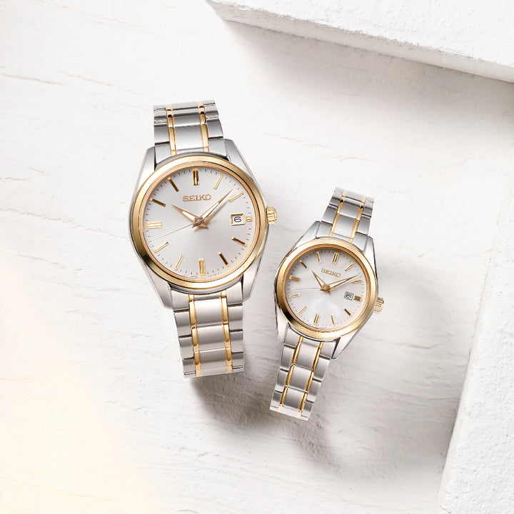 Twee Seiko dameshorloge SUR636P1 horloges met zilveren en gouden metalen banden en witte wijzerplaten worden gepresenteerd op een getextureerd wit oppervlak. Deze quartzhorloges hebben gouden uurmarkeringen, wijzers, kleine datumvensters en bieden 10ATM waterbestendigheid.