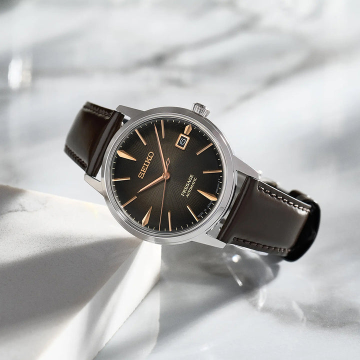 De Seiko Presage herenhorloge SRPJ17J1 heeft een bruine wijzerplaat, datumweergave, bruine leren band en zilveren kast. Dit automatische uurwerk staat op wit marmer, de elegantie wordt benadrukt door schaduwen op het Hardlex mineraalglas.