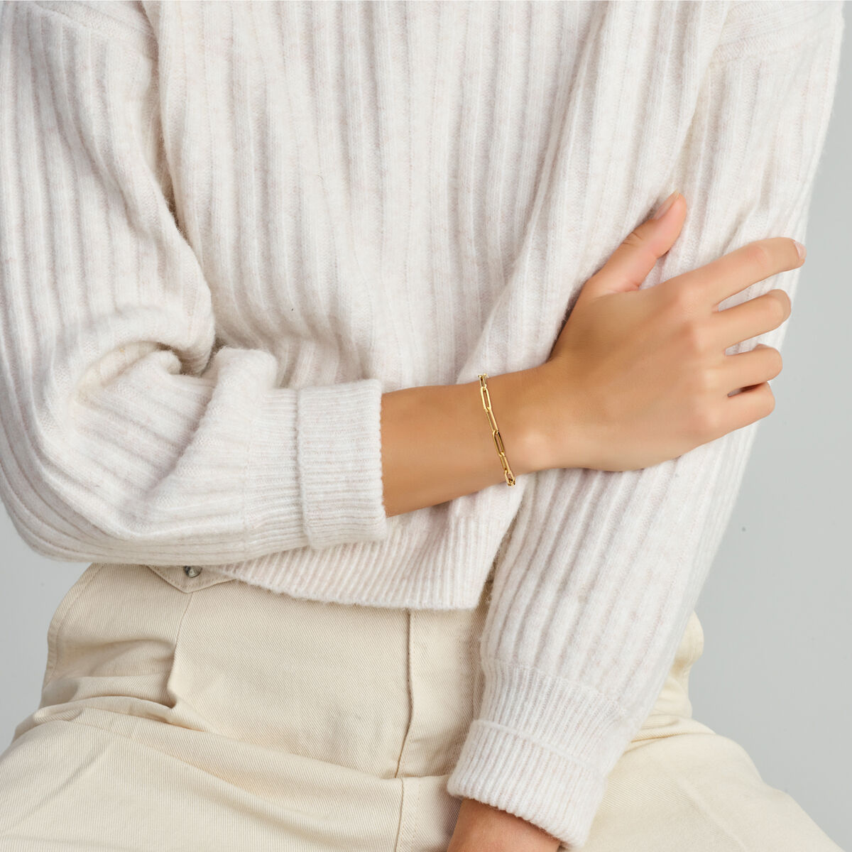 Een persoon in een geribde crèmekleurige trui met lange mouwen en een lichtbeige broek, zittend met één arm gekruist, showt een Van der Kooij Collectie Gouden armband dames paperclip vierkante buis met groot springslot 14K om zijn pols.
