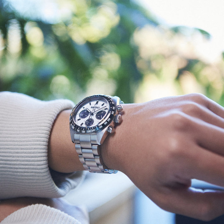 Een persoon draagt een Seiko Prospex SSC813P1 herenhorloge, met een zilveren chronograaf, saffierglas, witte wijzerplaat met meerdere wijzerplaten en metalen band. Op de wazige groene achtergrond dragen ze een licht, lang mouwen kledingstuk.