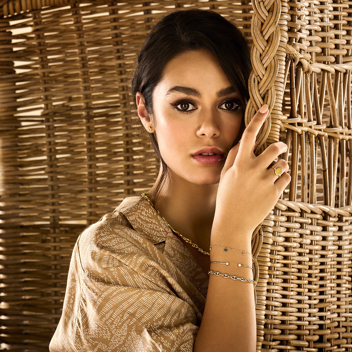 Een donkerharige vrouw poseert bij een rieten scherm, draagt een beige blouse met patroon, gouden sieraden en een Van der Kooij Collectie graveerring massief mat gediamanteerd 14K geelgoud terwijl ze naar de camera kijkt.