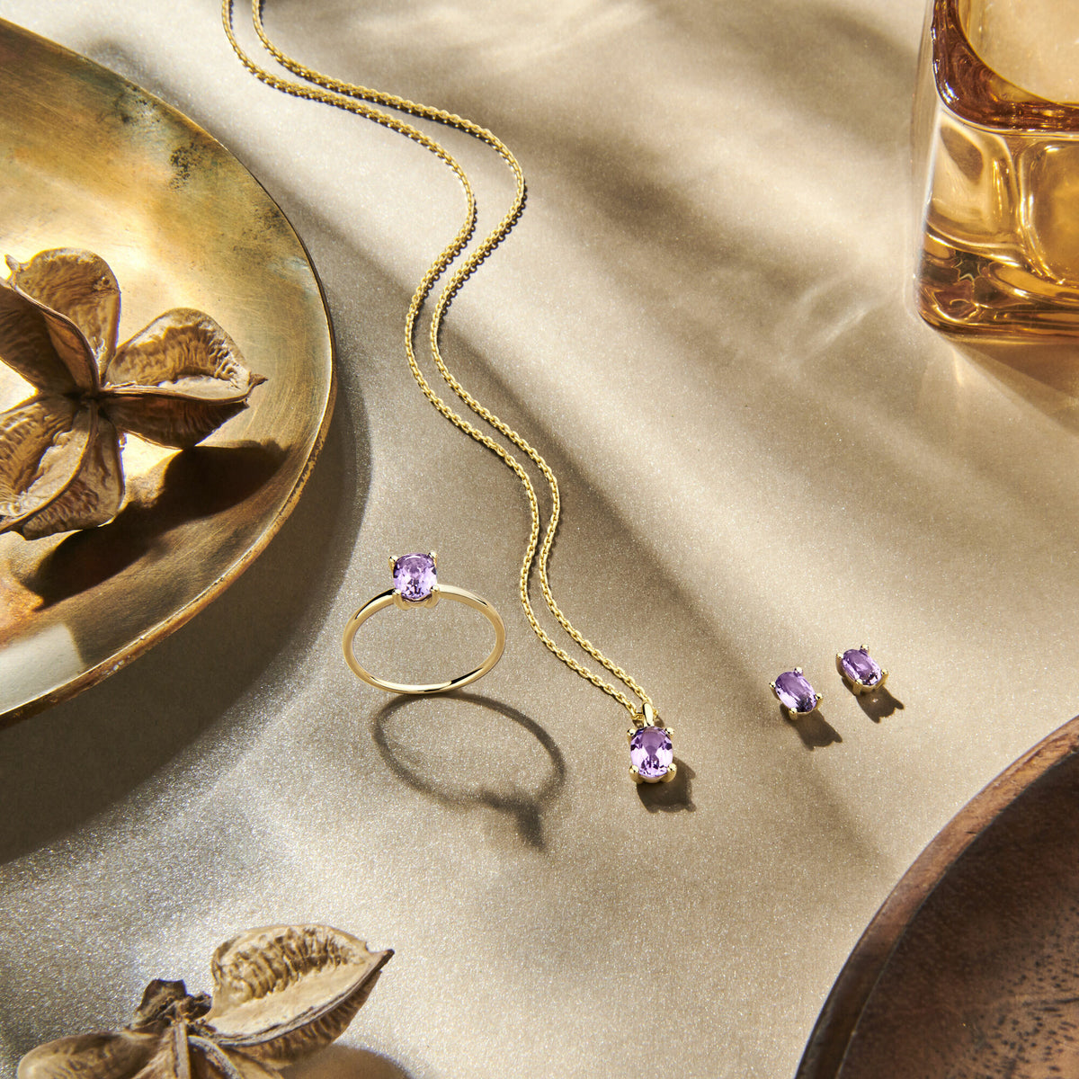 De Van der Kooij Collectie hanger amethist 14K geelgoud, samen met bijpassende ketting, ring en oorknopjes, staat elegant tentoongesteld op een metalen ondergrond naast kommen en gedroogde bloemen in warm licht.
