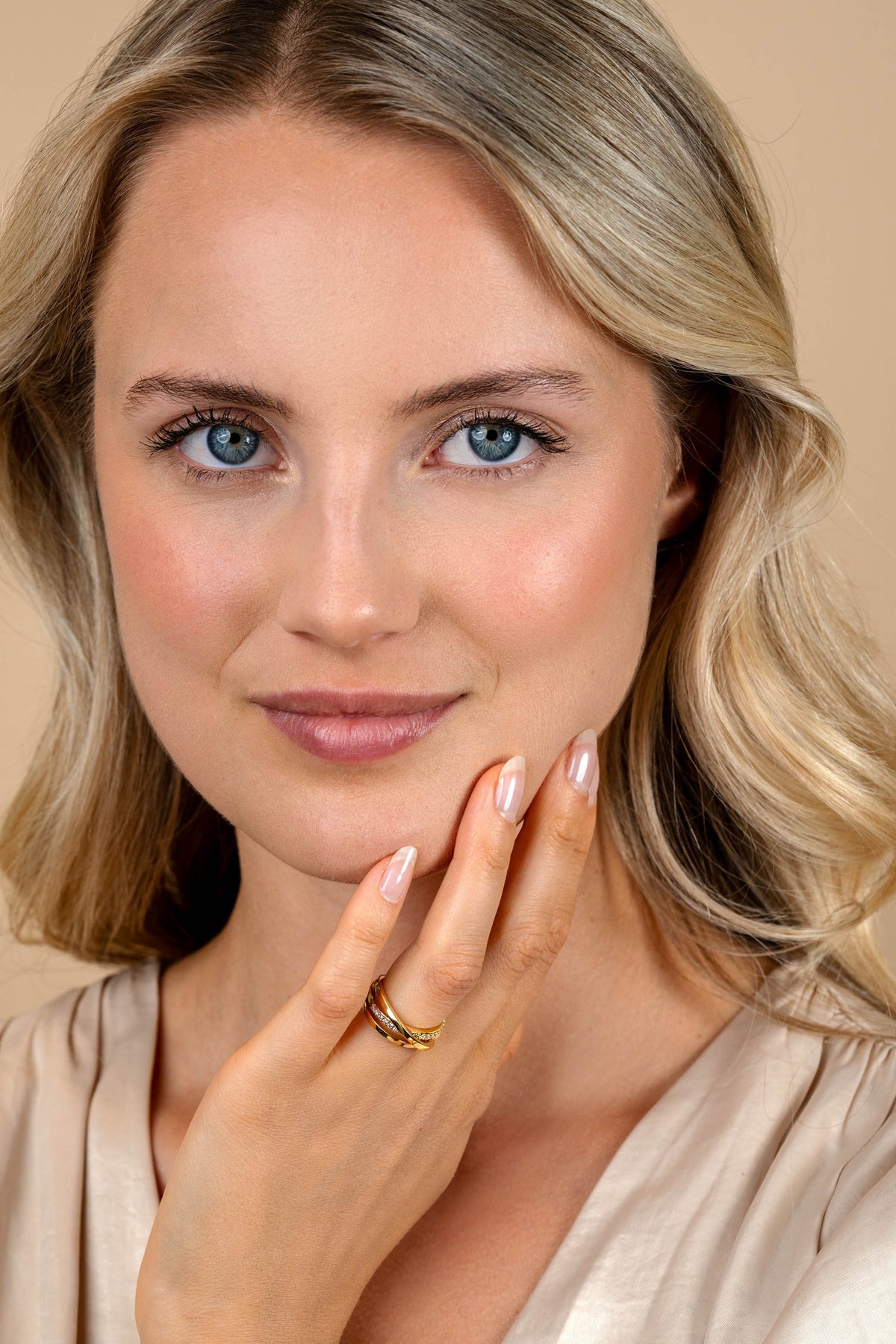 Een vrouw met golvend blond haar en blauwe ogen glimlacht zachtjes en legt haar gemanicuurde hand versierd met de Zinzi Ring Gold Plated ZIR1774Y op haar kin. Ze draagt een lichtbeige topje en natuurlijke make-up tegen een beige achtergrond.