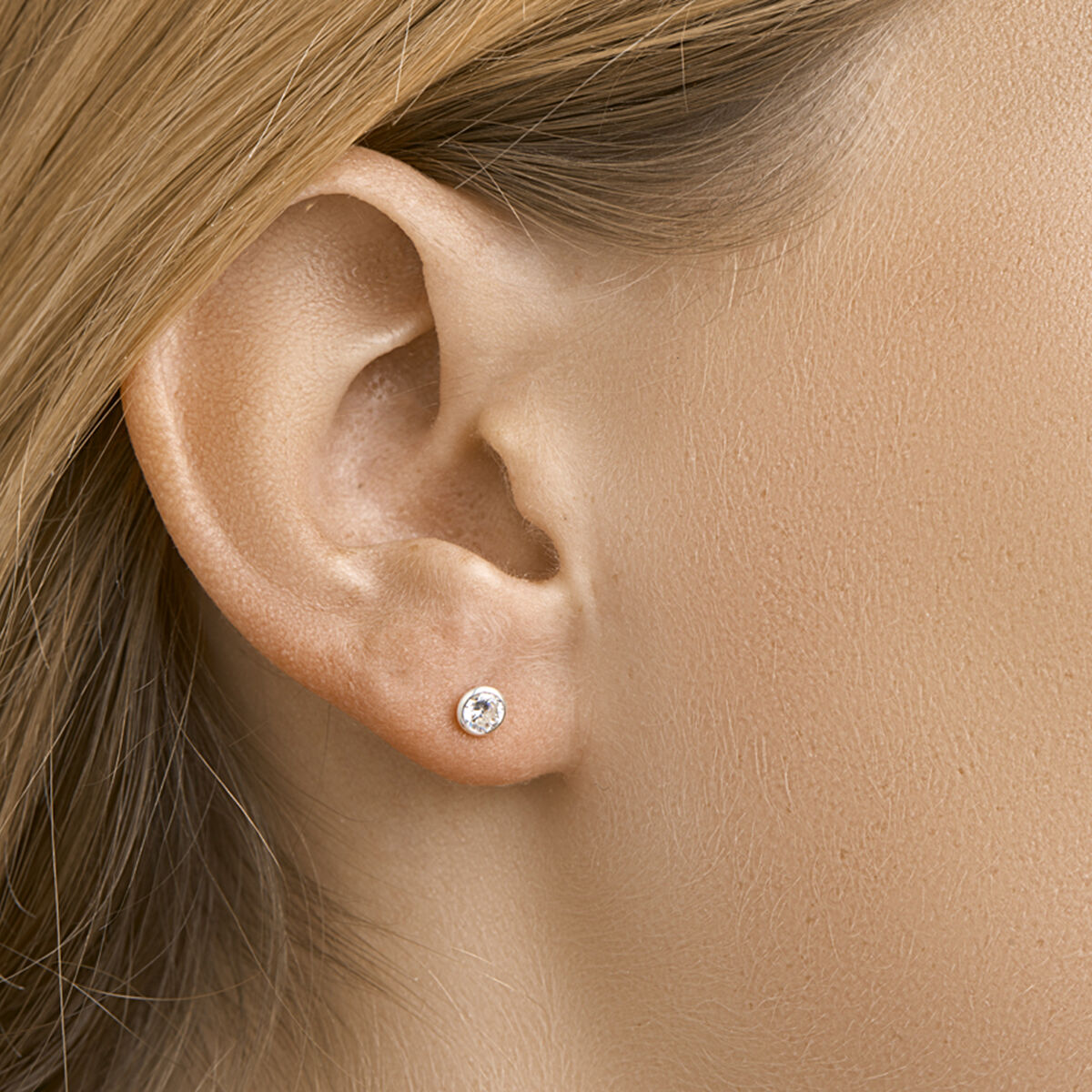 Close-up van een vrouwenoor met steil lichtbruin haar, met oorknoppen zirkonia zilver wit uit de Van der Kooij Collectie - kleine ronde oorstekers met een witte zirkonia steen in een zilveren zetting.
