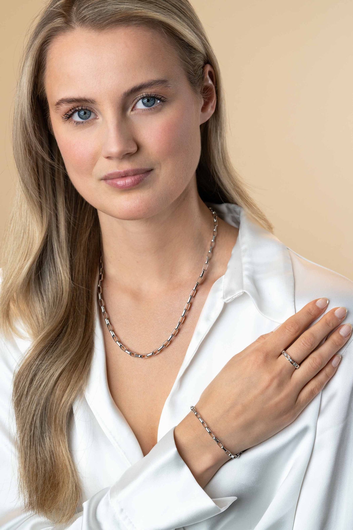 Een vrouw met lang blond haar draagt een witte blouse en zilveren Zinzi-sieraden, waaronder de Zinzi Armband zilver ZIA2514. Ze kijkt met een vriendelijke glimlach in de camera, poserend tegen een beige achtergrond.