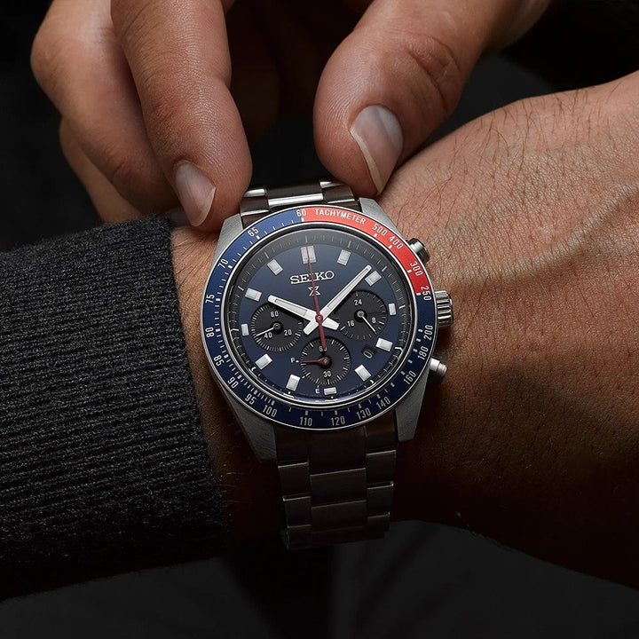 Een persoon draagt een Seiko Prospex SSC913P1 chronograafhorloge van roestvrij staal, met een zwarte wijzerplaat, blauwrode lunette, subdials en tachymetermarkeringen. Ze passen het op zonne-energie werkende uurwerk op hun pols aan.