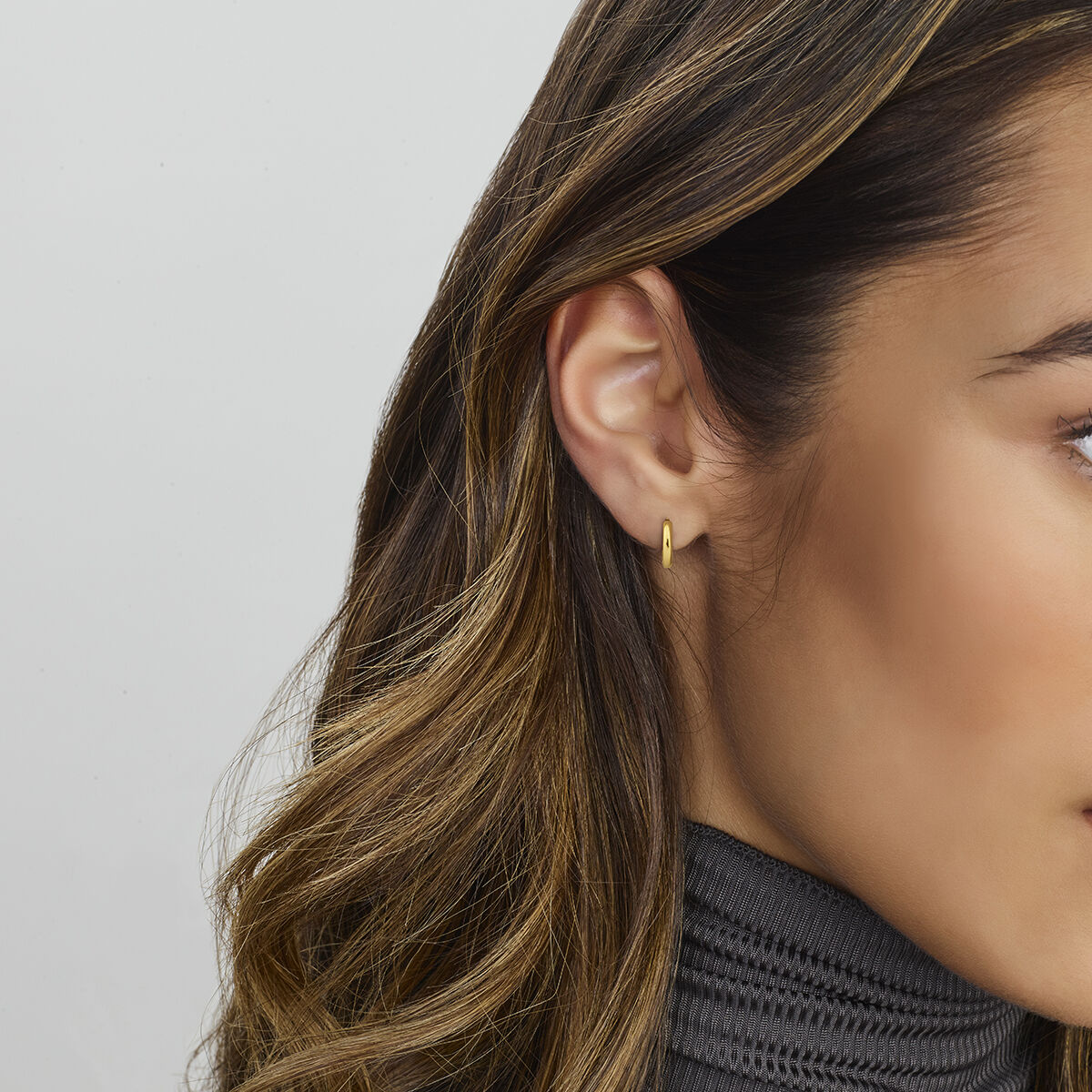 Close-up van het rechteroor van een vrouw met Van der Kooij Collectie klapoorringen 14K geelgoud met scharniersluiting. Ze heeft lang, golvend bruin haar, een grijze geribbelde coltrui en de achtergrond is effen en lichtgekleurd.