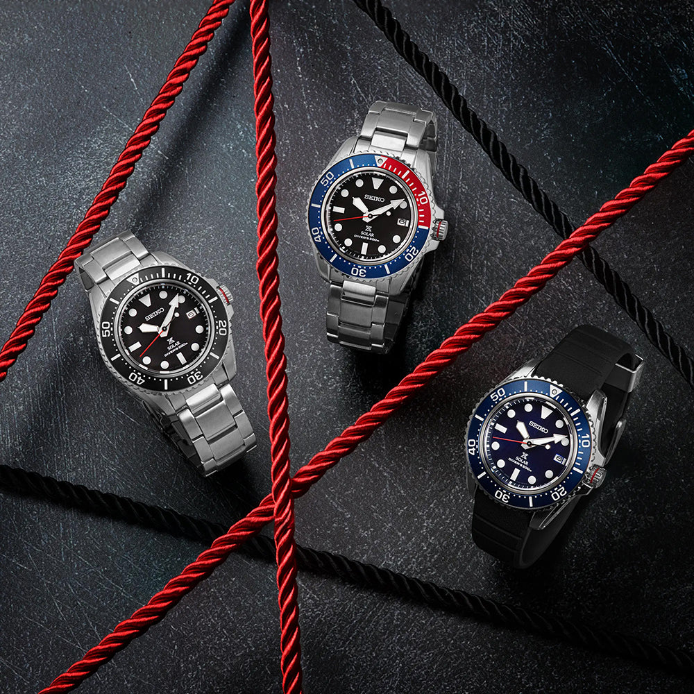Drie duikhorloges met draaibare bezels en metalen bandjes, waaronder een Seiko Prospex heren horloge SNE589P1 met solar uurwerk, zijn diagonaal gerangschikt op een getextureerd donker oppervlak. Er is een rood touw overheen gelegd, wat een visueel opvallend contrast creëert met hun metaalachtige tinten.