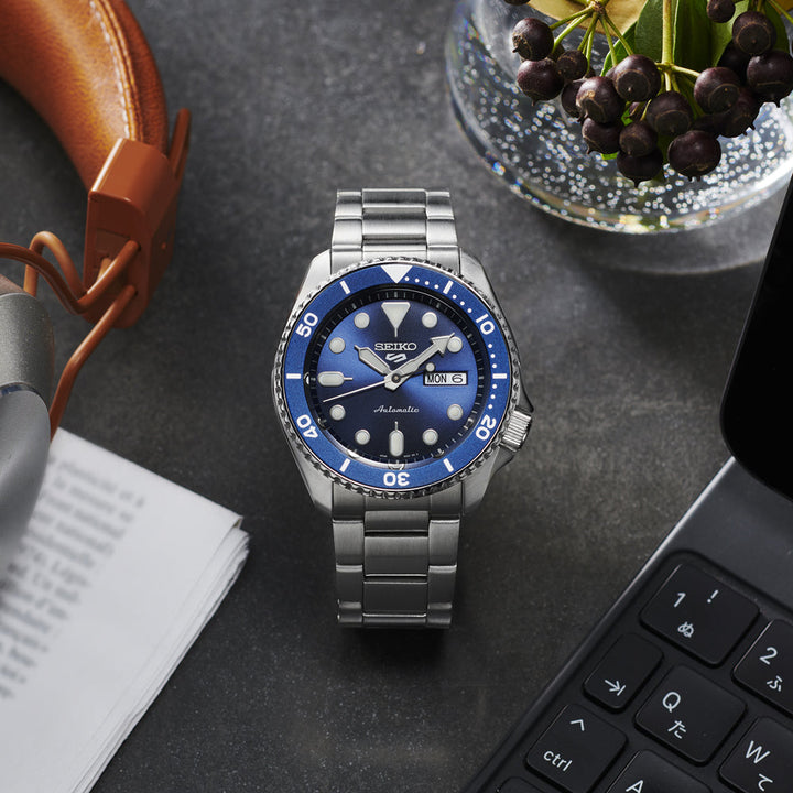 Naast een laptop, koptelefoon, papieren en een schaal met bessen staat het Seiko 5 Sports heren horloge SRPD51K1. Dit zilveren horloge van Seiko heeft een blauwe wijzerplaat die elegant de dag en datum weergeeft.