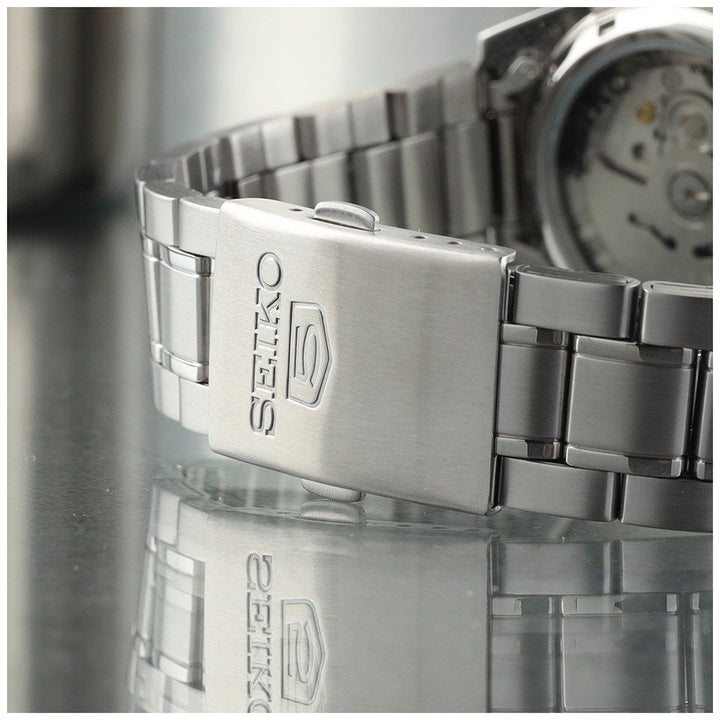 Een close-up van het Seiko heren horloge SNKL45K1 horlogebandje met een vouwsluiting van roestvrij staal, met prominent de gravure Seiko. Op de achtergrond is op de transparante achterkant een automatisch uurwerk te zien.