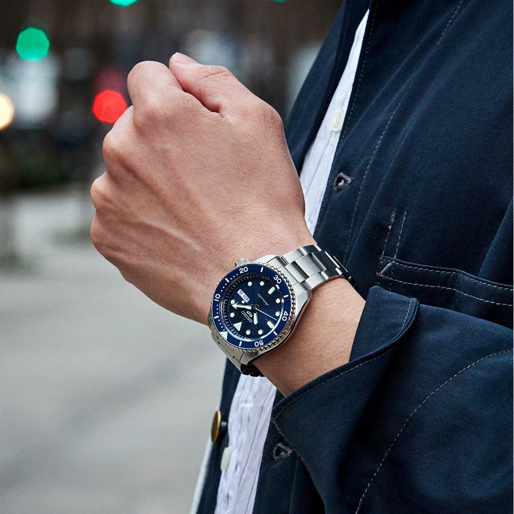 Een persoon in een donker jasje en wit shirt toont de Seiko 5 Sports heren horloge SRPD51K1, een elegant zilveren horloge met een blauwe wijzerplaat en bezel. Ze zijn buiten met wazige straatverlichting die de achtergrond versterkt.