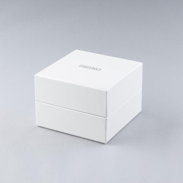 Een witte Seiko horlogedoos met het logo van het merk hint naar de elegantie van het Seiko heren horloge SUR527P1 met saffierglas en 10ATM waterdichtheid. Het minimalistische ontwerp staat tegen een grijze achtergrond, weergegeven in een lichte hoek.