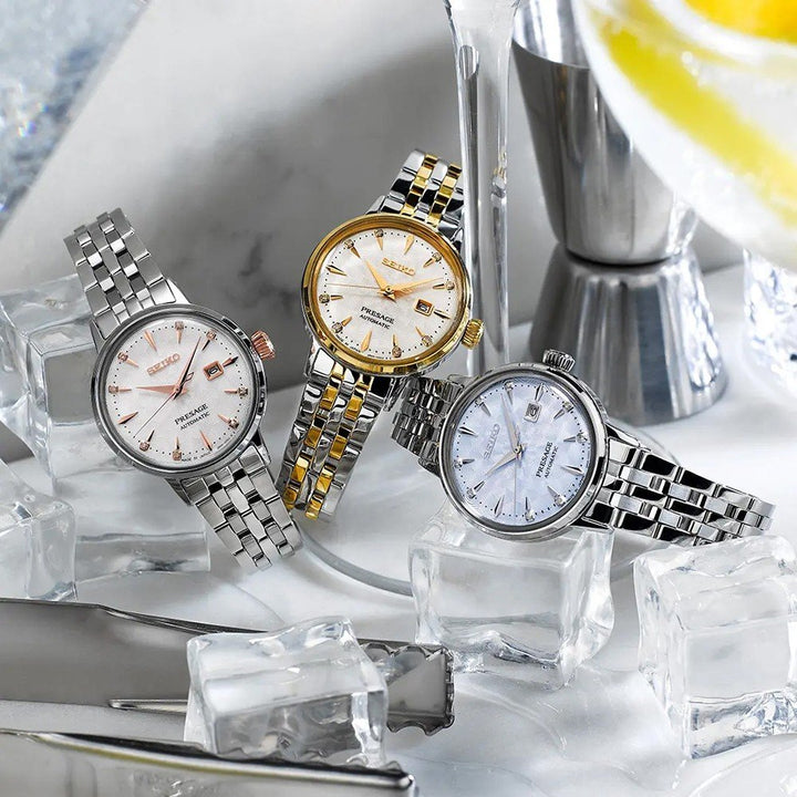 Drie elegante polshorloges met metalen banden rusten op ijzige kubussen, elk met unieke bezels: zilver, goud en goud-geaccentueerd. Het Seiko Presage dameshorloge SRE009J1 voegt verfijning toe met zijn Hardlex mineraalglass voorkant. Een cocktailshaker en een schijfje citroen verschijnen op de achtergrond.
