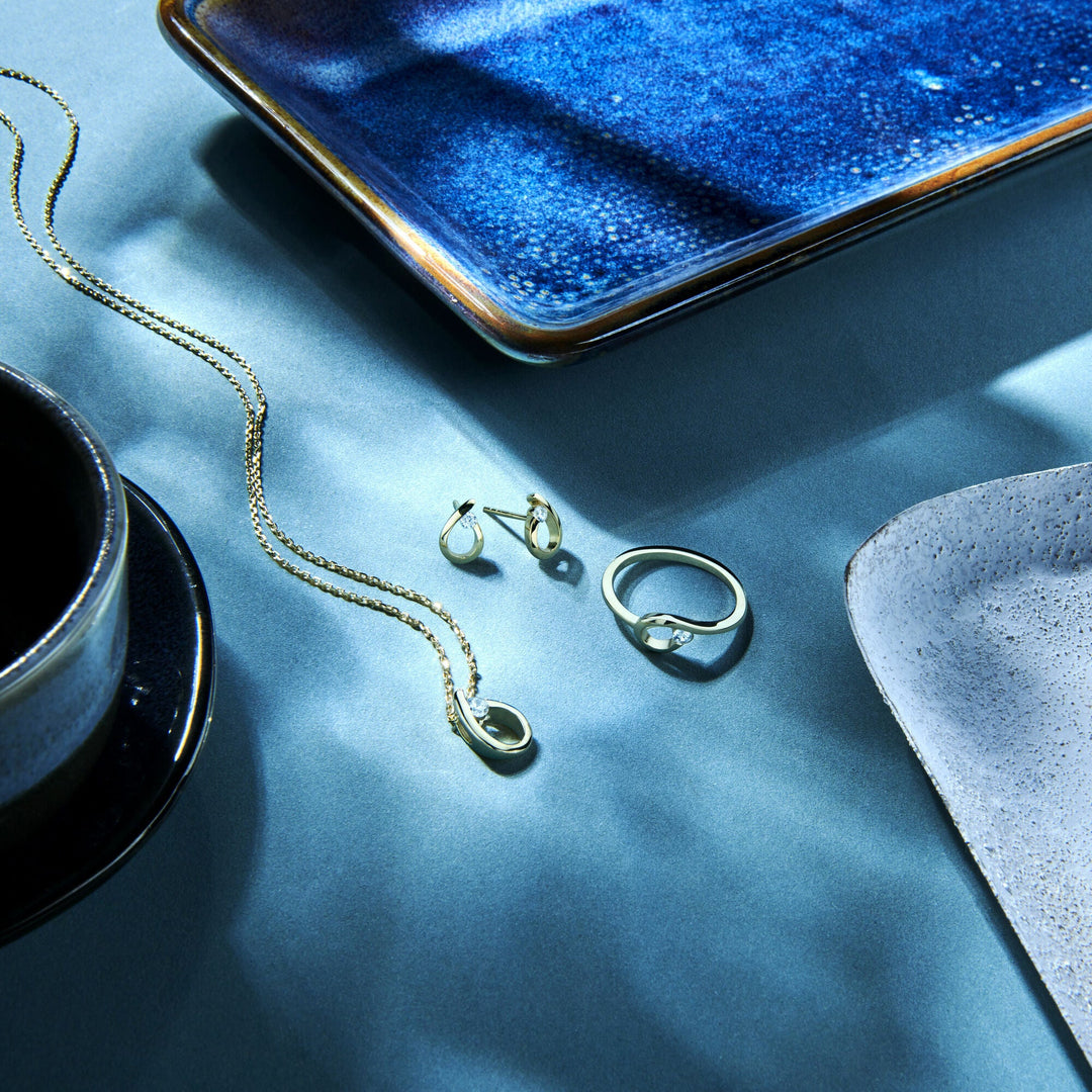 Gouden sieraden uit de Van der Kooij Collectie, met onder andere een ring met zirkonia in 14 karaat geelgoud, een ketting en oorbellen, gepresenteerd op een blauwe ondergrond met keramische schalen in zacht, elegant licht.