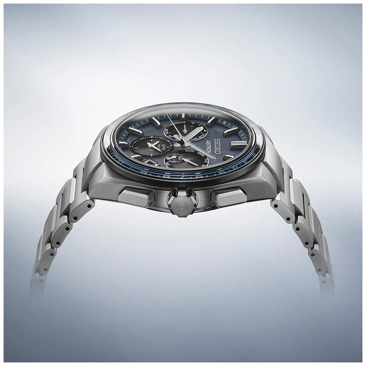Zijaanzicht van het gestroomlijnde Seiko Astron SSH109J1 herenhorloge, met een zilveren design, donkerblauwe wijzerplaat met subdials en tachymeter. Het beschikt over saffierglas en metaalachtige glans tegen een zachte gradiëntachtergrond, wat de verfijnde stijl benadrukt.