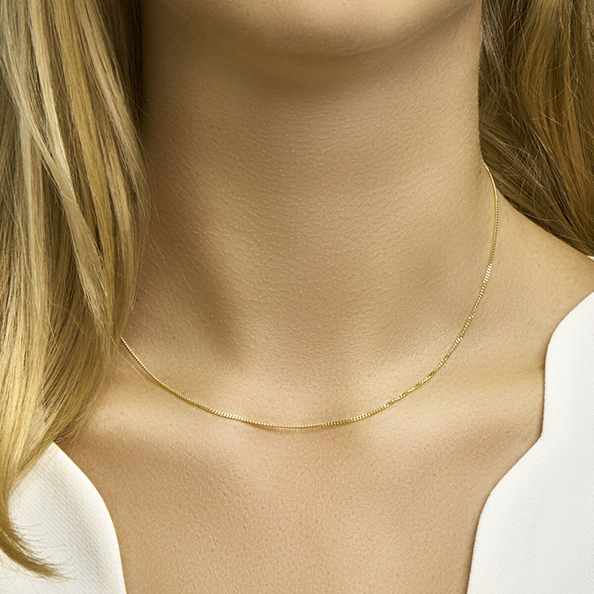Een close-up van een vrouw met een lichte huid en blond haar in een witte top, waarbij haar nek benadrukt wordt, versierd met de Van der Kooij Collectie collier gourmette 4-zijdes geslepen 1,2 mm in 14K geelgoud.