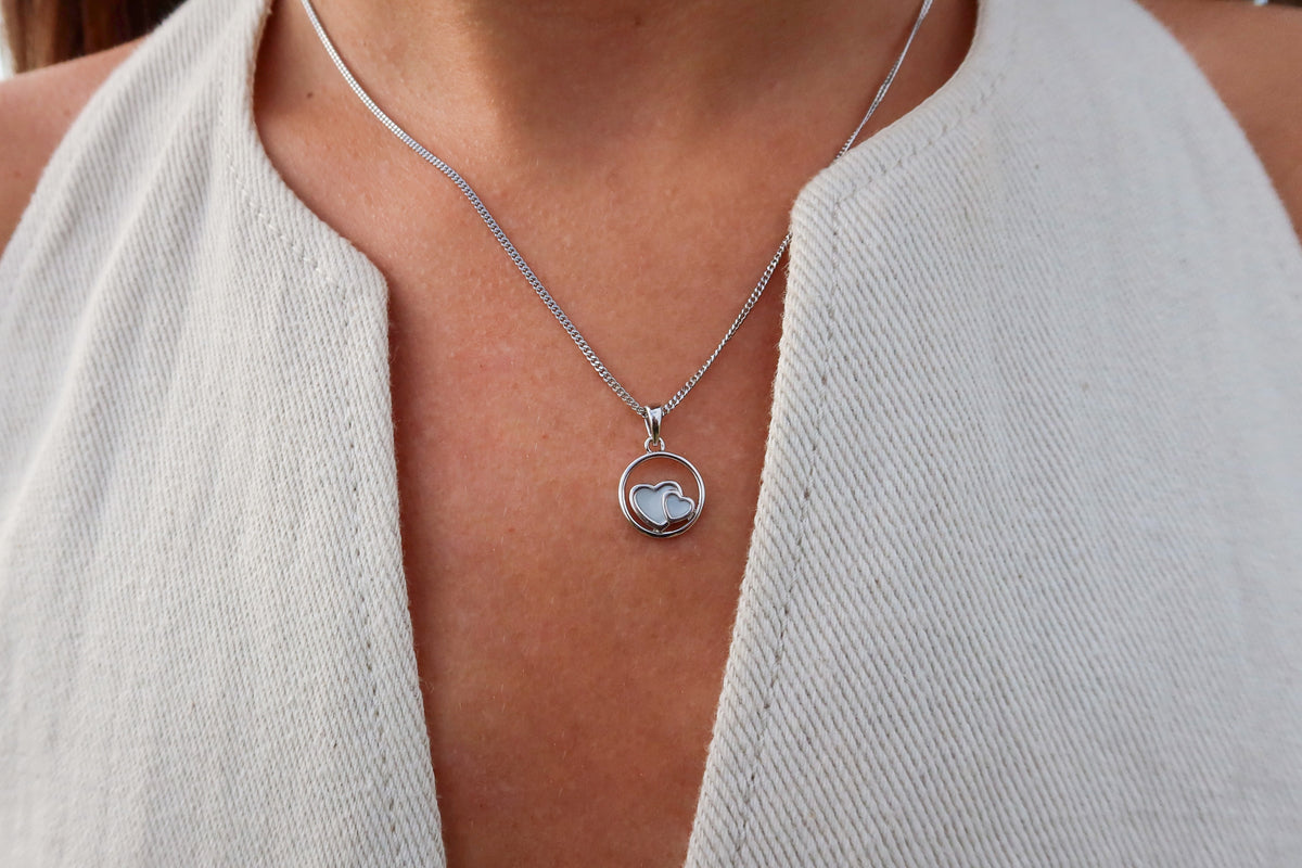 Een persoon draagt een mouwloze top met crèmekleurige textuur en de Amélie AsHanger massief halsketting van Juwelier van der Kooij, met een ronde hanger met hartmotief. De focus ligt op de ketting en de bovenkant van de borst.
