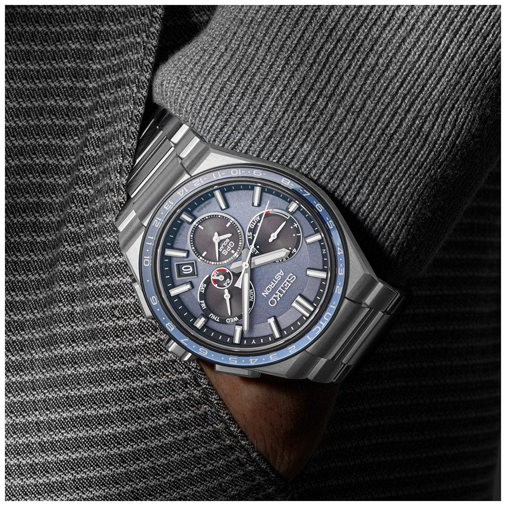 De Seiko Astron SSH109J1 herenhorloge heeft een donkergrijze wijzerplaat met meerdere wijzerplaten, waaronder een datumweergave en chronograaf, beschermd door robuust saffierglas. Gestyled met een getextureerd grijs pak, rust de hand van de drager in de zak.