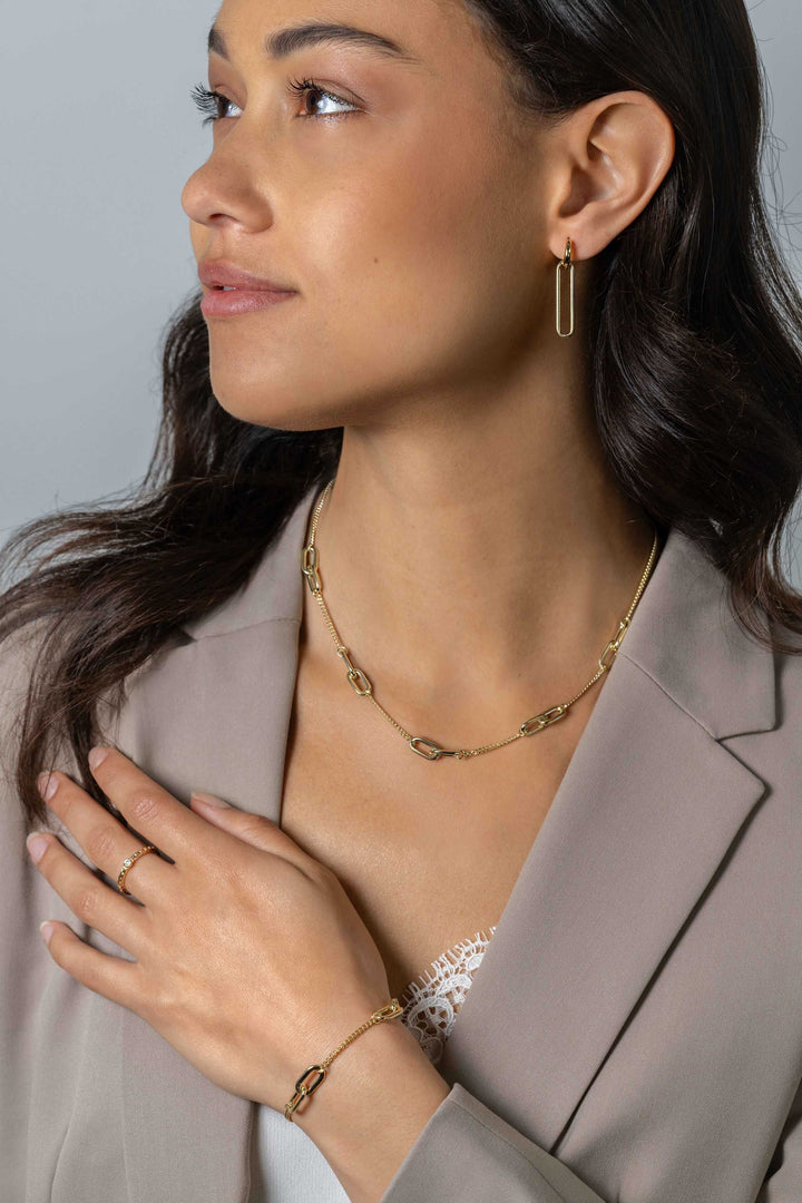 Een vrouw in een lichtbeige blazer en wit kanten topje showt Zinzi Oorbedels Gold Plated Zich2416 G sieraden van Zinzi, waaronder een kettingketting, hoepeloorbellen, een ovaalvormige armband en een ring terwijl ze naar links kijkt.