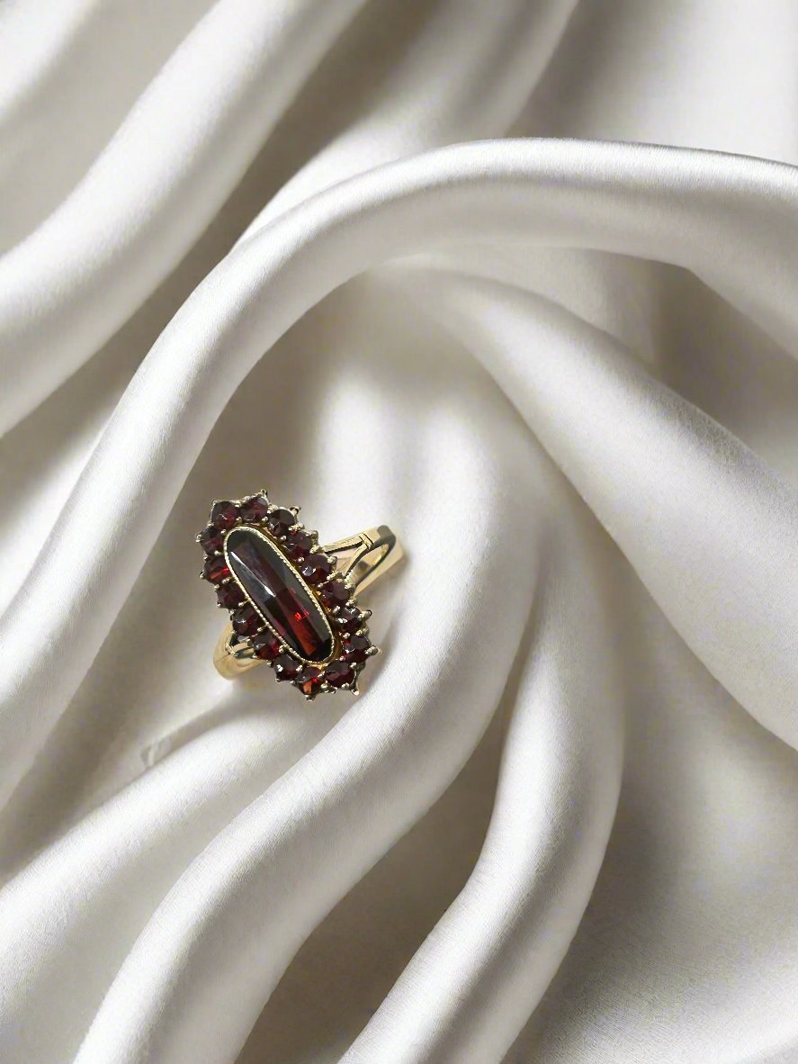 De Vintage Collectie ring van Juwelier van der Kooij, een 14 karaat geelgouden creatie met een ovale granaat van 3,7 gram en omringd door kleinere donkerrode stenen, contrasteert elegant met de zachte witte zijden stof waarop hij rust.