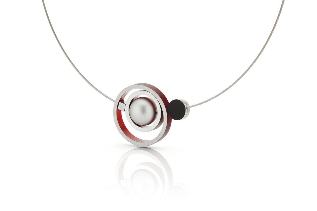 De Collier Ringen in elkaar Rood C187R van CLIC Creations heeft een minimalistisch ontwerp met een dunne draad en een grote zilveren bol omringd door rode en zwarte ringen, wat een moderne touch biedt. Handgemaakt in Nederland, straalt het elegantie uit tegen een effen witte achtergrond.