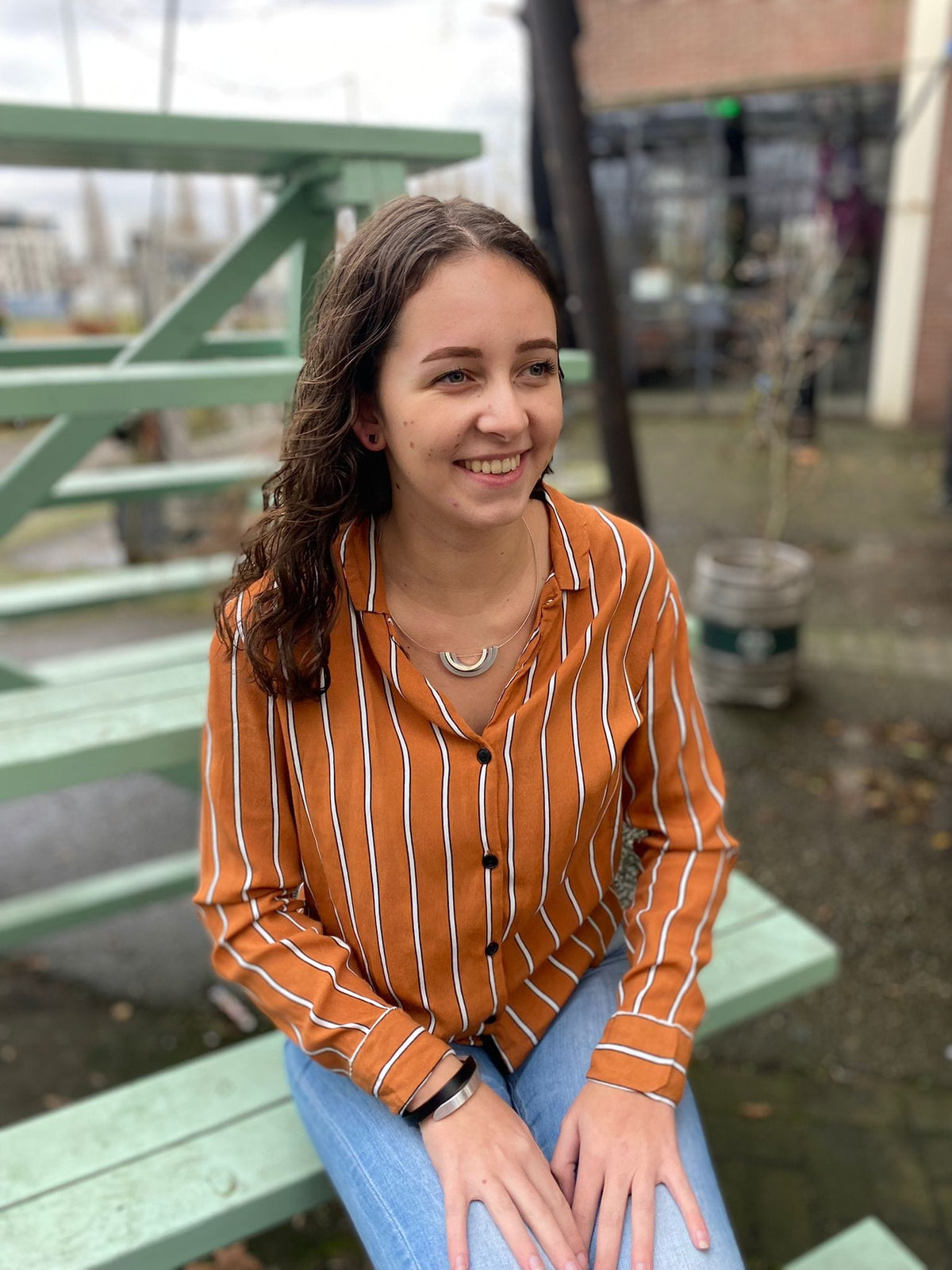 Een vrouw met lang bruin haar zit op een mintgroene trap, met een unieke glimlach. Ze draagt een bruin-wit gestreept shirt en een blauwe spijkerbroek, vlak bij een gebouw met grote ramen en een buitenruimte. Haar moeiteloos chique stijl lijkt op de handgemaakte Armband Cs beschouwd Zwart A1Z van CLIC Creations.