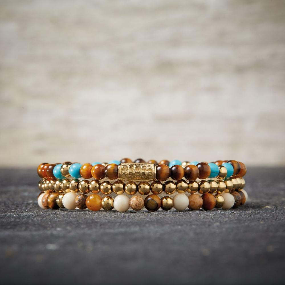 Een stapeltje van vier kralenarmbanden, waaronder de Rebel & Rose Mix Tiger Turquoise 18 kt geelgoud geïoniseerd 4mm kralen armband dames, heeft gouden, bruine, crème, turquoise en tan kralen op een donker oppervlak met een licht vervaagde achtergrond-perfecte dames sieraden.
