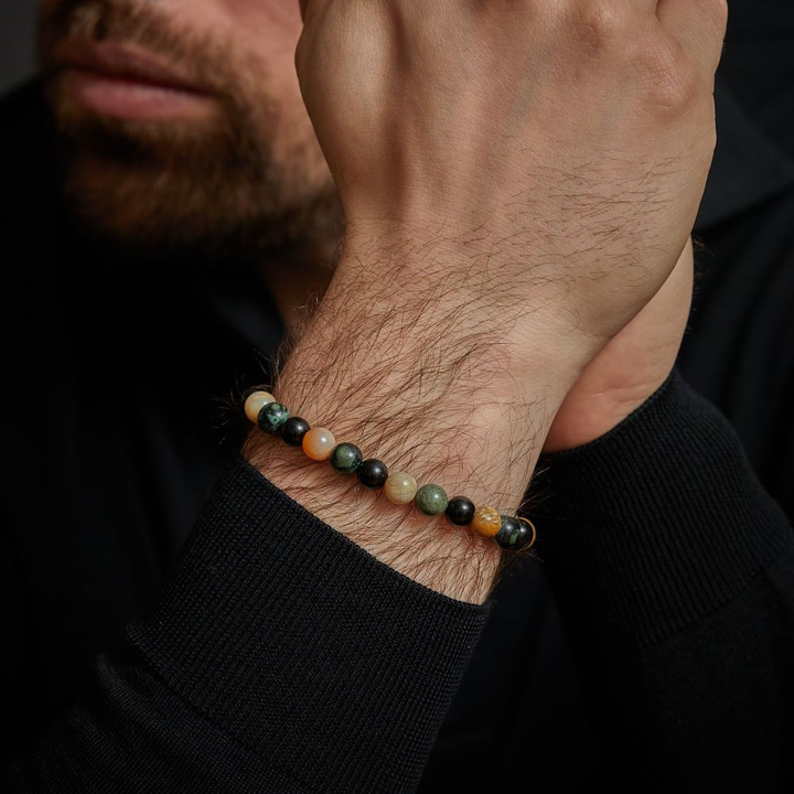 Een close-up van de handen van een bebaard persoon, die de Rebel & Rose Autumn Storm - 8mm Kralen Heren Armband met groene, zwarte en bruine stenen draagt, tegen een donkere achtergrond. De persoon is gekleed in een zwart shirt met lange mouwen.