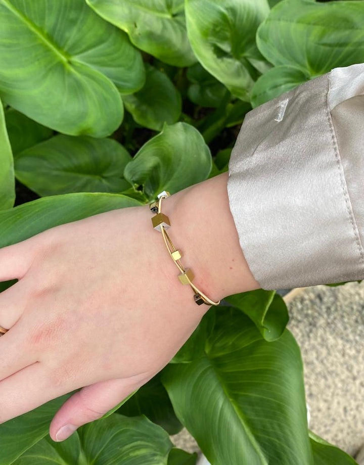 Een persoon die de CLIC Creations Armband speelse kubusjes goud A257G draagt, laat zijn hand rusten op weelderige groene bladeren. Ze zijn elegant gestyled in een lichtgekleurd satijnen shirt met geometrische gouden accenten.