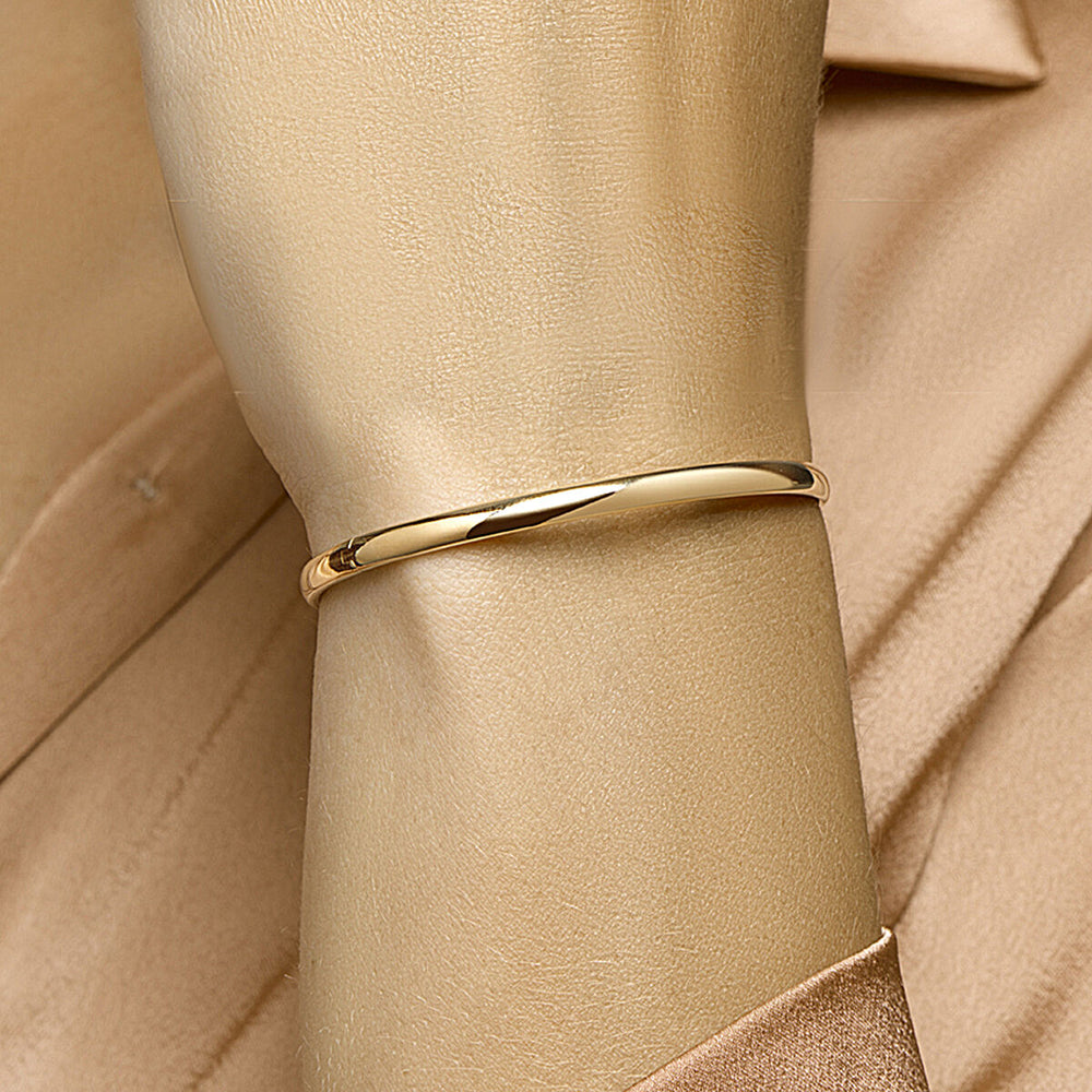 Een close-up van een Van der Kooij Collectie bangle scharnier massief ovale buis 4,0 mm Zilgold (geelgoud met zilverkern) gedragen om de pols over een beige satijnen shirt, met bijpassende achtergrond die het glanzende, eenvoudige ontwerp laat zien.
