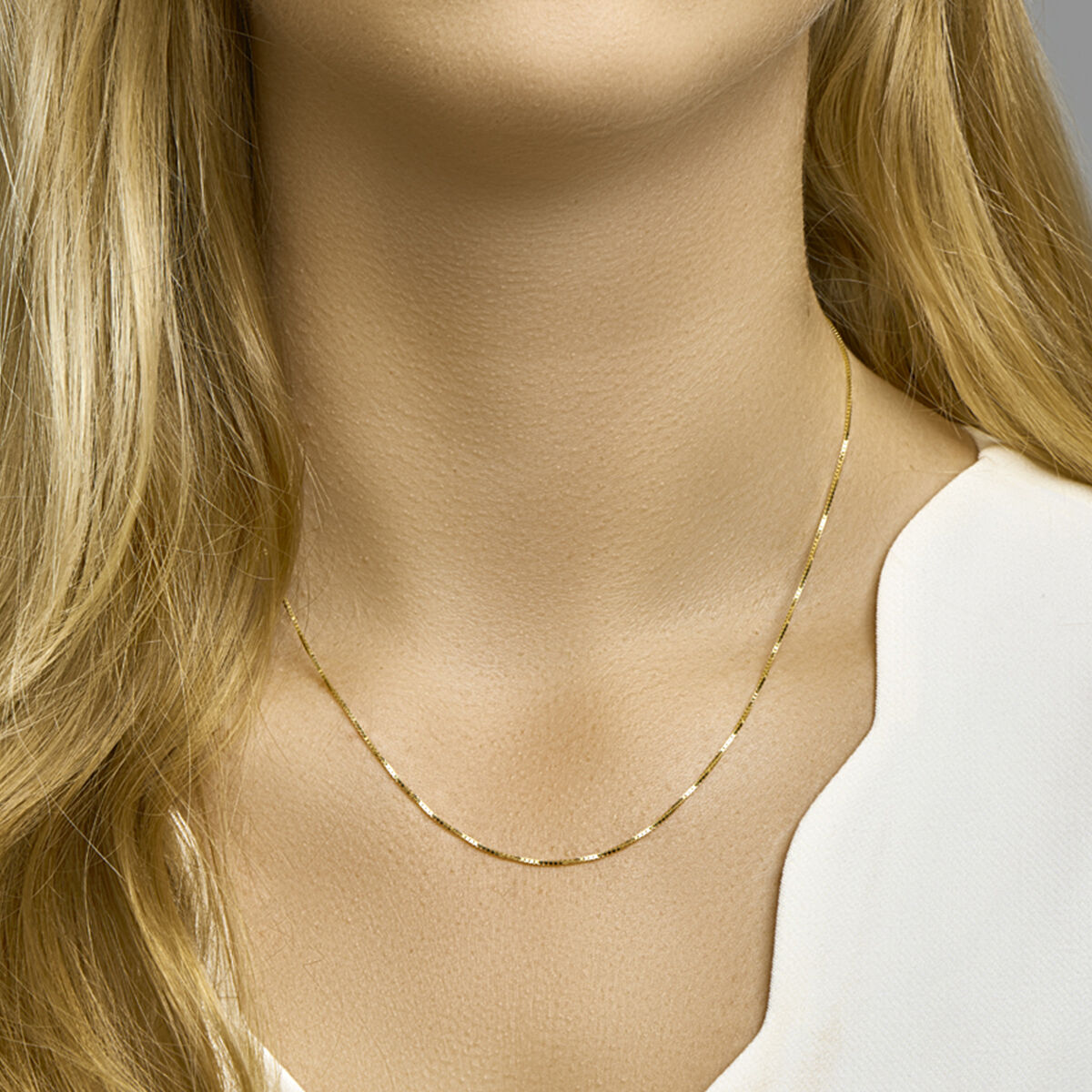 Een vrouw met lang blond haar draagt een Van der Kooij Collectie Venetiaans collier 0,8 mm 14K geelgoud, gecombineerd met een wit topje met inkeping. De focus ligt op de ketting, die haar nek en schouders accentueert.