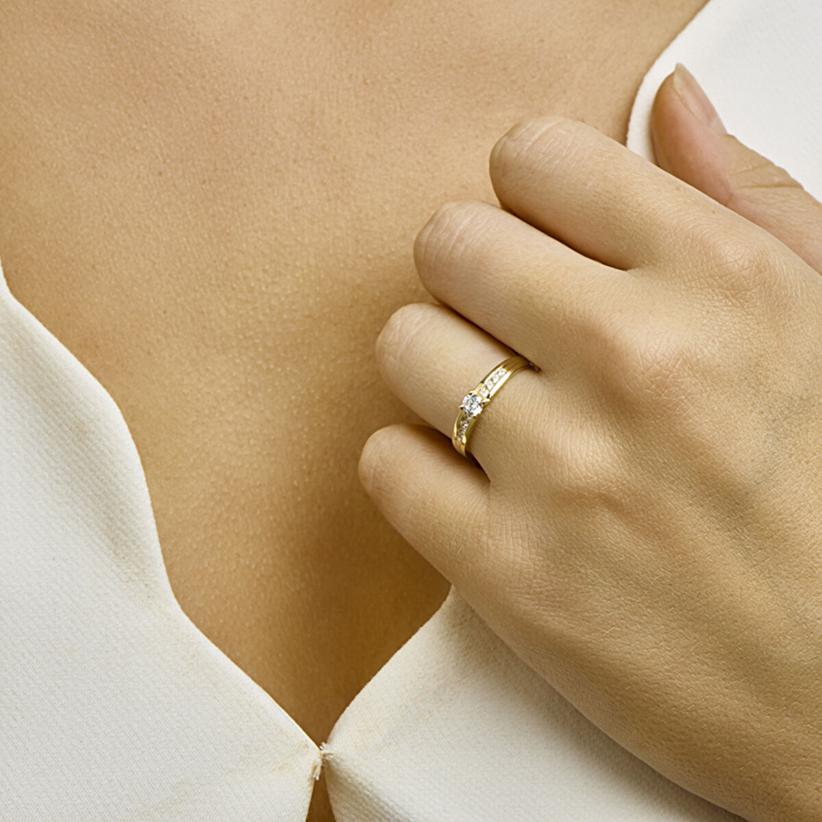 Een close-up van de hand van een vrouw die de Van der Kooij Collectie ring zirkonia 14K geelgoud draagt, rustend op haar borst boven een wit kledingstuk met v-hals.