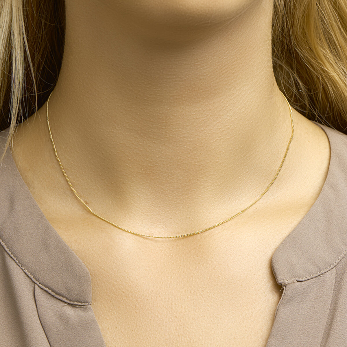 Een close-up van de hals van een vrouw met het Van der Kooij Collectie collier gourmette 1,0 mm 14K geelgoud, gecombineerd met een gedeeltelijk zichtbare taupe blouse.