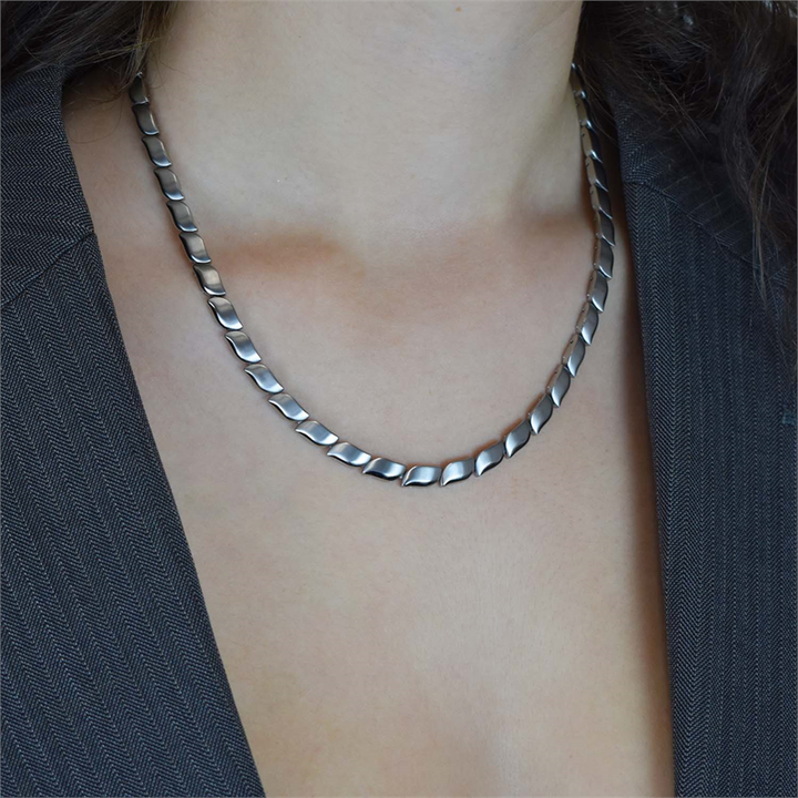 Een persoon draagt de elegante Boccia Titanium Collier 08038-01, een zilverkleurige damesketting met een visgraatmotief, gecombineerd met een donkere, getextureerde blazer. De platliggende ketting contrasteert prachtig tegen hun huid in een neutrale, soft-focus achtergrond.