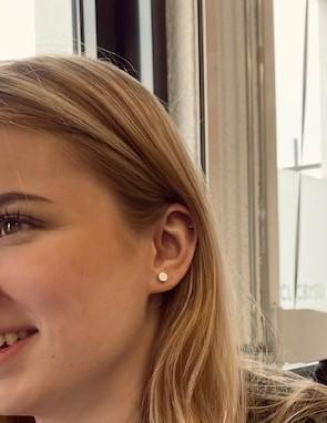 Een glimlachende persoon met lang blond haar en een kleine, handgemaakte oorbel zit bij een raam, tegen de achtergrond van een stadsgezicht. De elegante Oorsteker Klein rond Grijs O36AG van CLIC Creations is hypoallergeen en handgemaakt in Nederland.
