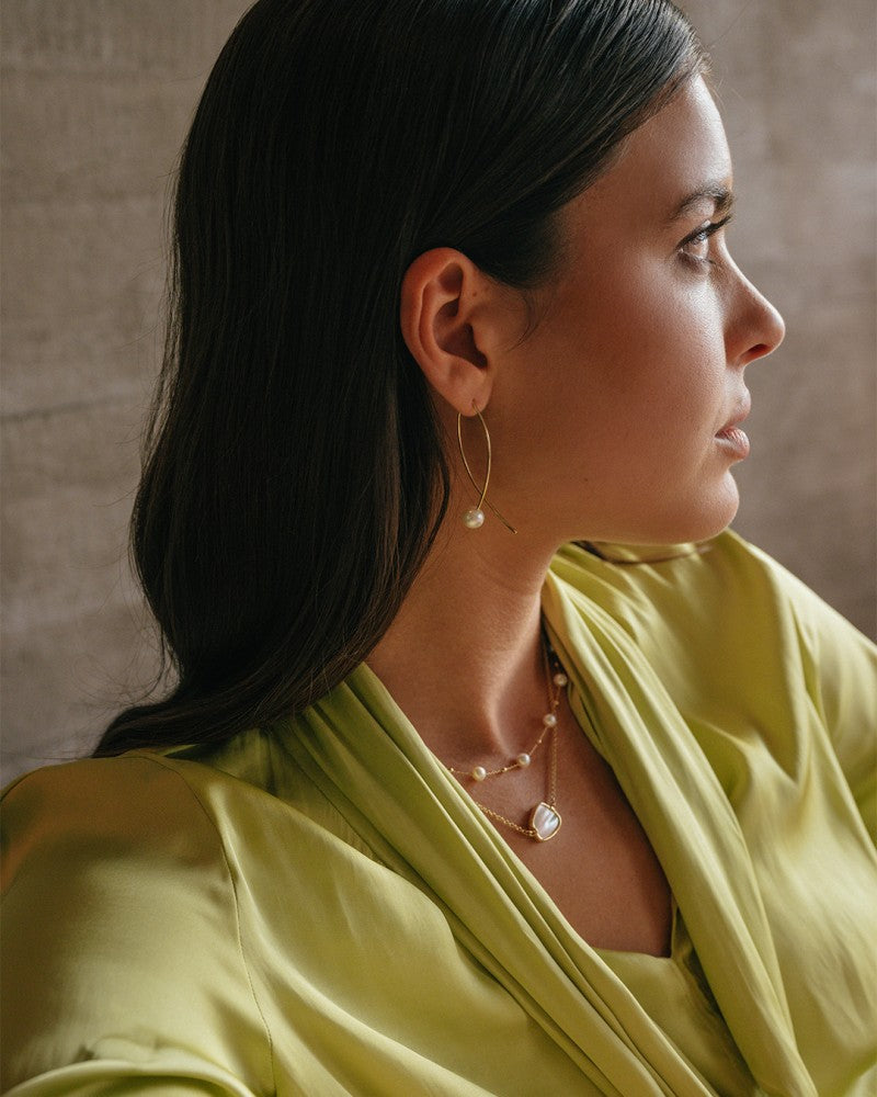 Een vrouw met lang bruin haar in een lichtgroene satijnen blouse zit in profiel, draagt Opvallende Geelgouden Oorbellen met Grote Parel 14 Karaat van Excellent Jewelry en gelaagde gouden kettingen met kleine hangers, en staart peinzend voor zich uit.